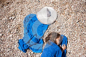 Beautiful little girl lying on beach wrapped in blue towel near white hat