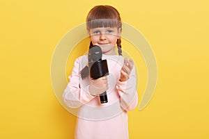 Beautiful little girl with microphone performing, looking at camera with charming smile, raising hand, looks a bit shy, child