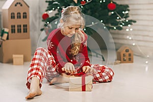 Beautiful little girl opens a gift at home under a kraft paper tree tied with a red ribbon, new year mood. Holiday concept for