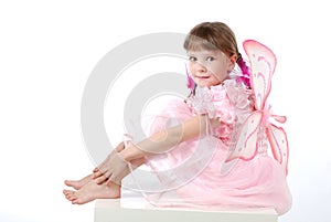 Beautiful little girl in a pink dress with butterfly wings is sitting on a table on a white background