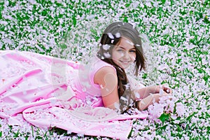 Beautiful little girl in pink dress with smiling happy face lying on green grass covered with spring flower blossom