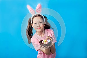 Beautiful little girl in a pink t-shirt with bunny ears holding a nest with easter eggs on a blue background