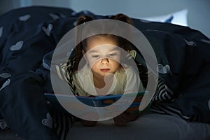 Beautiful little girl reading book in bed at night.