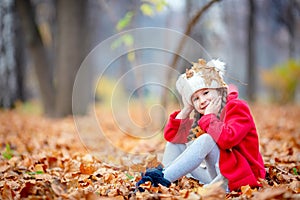 Beautiful little girl in red coat sitting on autumn leaves with falling foliage outdoors in a park