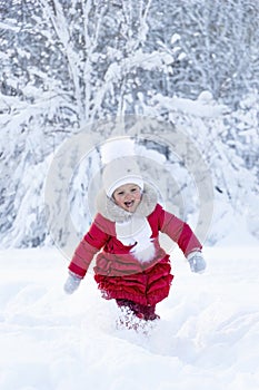 A beautiful little girl in a red coat and white hat with a scarf runs along a snowy path in a winter park. Smile and