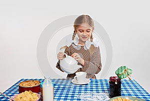 Beautiful little girl in retro clothes pouring tea into cup, serving breakfast against grey studio background. Concept