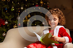 Beautiful little girl sits on the chair next to a nicely decorated Christmas tree