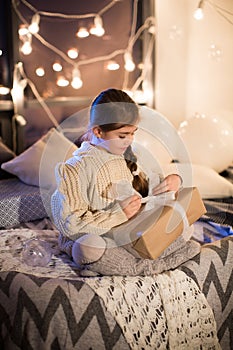 Beautiful little girl sitting on bed with a gift in its hands. Deploy Christmas gift box on holiday in the evening in