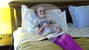 Beautiful little girl sitting on the bed in the hotel and watching TV. In the hands of a girl holding a remote control