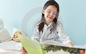 Beautiful little girl sitting on the bed and reading the fairy tale book. Kid wearing pajamas relaxing and reading interesting