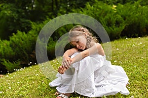 A beautiful little girl sitting on the bright green grass and embracing her knees with her