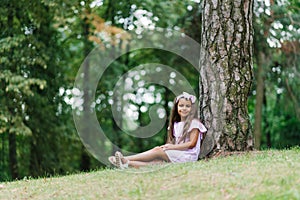 A beautiful little girl sitting near a tree, relaxing, enjoying a sunny day in a green park. Summer school holidays