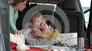 Beautiful little girl sitting in an open trunk of a car on the river bank of the sea playing on a yellow ukulele