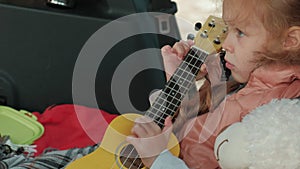 Beautiful little girl sitting in an open trunk of a car on the river bank of the sea playing on a yellow ukulele
