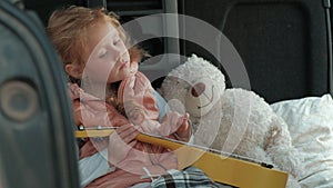 Beautiful little girl sitting in an open trunk of a car on the river bank of the sea playing on a yellow ukulele