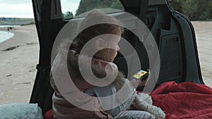 Beautiful little girl sitting in an open trunk of a car on the river bank of the sea uses a smartphone