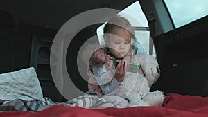 Beautiful little girl sitting in an open trunk of a car on the river bank of the sea uses a smartphone