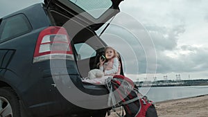 Beautiful little girl sitting in an open trunk of a car on the river bank of the sea uses a smartphone