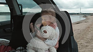 Beautiful little girl sitting in an open trunk of a car on the river bank of the sea uses a smartphone