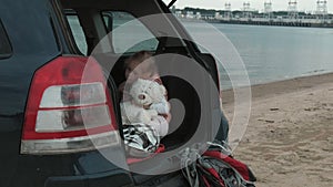 Beautiful little girl sitting in an open trunk of a car on the river bank of the sea uses a smartphone