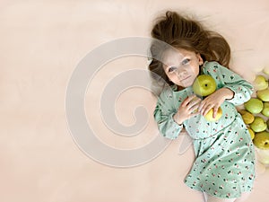 A beautiful little girl smiles and holds apples in her hands. Green fruit. The child lies on the bed and looks at the camera