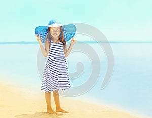 Beautiful little girl in a striped dress and summer straw hat relaxing on the beach near sea
