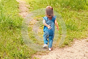 A beautiful little girl walks along a forest path. The baby is walking along a sandy path among the grass. A child runs away from