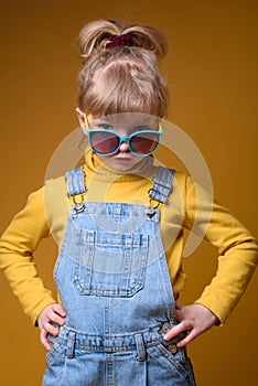 Beautiful little girl wearing stylish denim overall, yellow roll neck jumper and colorful sunglasses