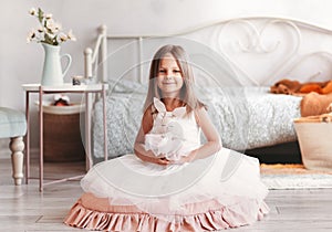 Beautiful little girl in white dress sits on the floor in a light room. Looking at camera. Childhood