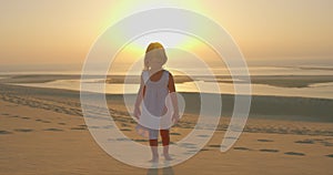 A beautiful little girl in a white dress walks barefoot on the sand at a golden sunset against the backdrop of the ocean