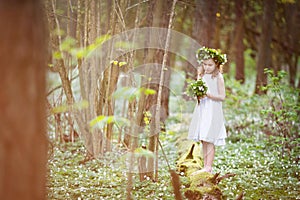 Beautiful  little girl in a white dress walks in the spring wood. A portrait of the pretty little girl with a wreath from spring