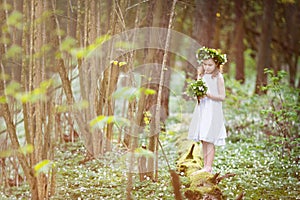 Beautiful  little girl in a white dress walks in the spring wood. A portrait of the pretty little girl with a wreath from spring