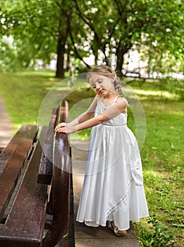 A beautiful little girl in a white long dress staying near the bench in the park with her