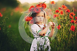Beautiful little girl with a wreath of poppies in a white dress and collects a bouquet of wildflowers. cute child in poppies field