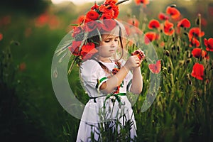 Beautiful little girl with a wreath of poppies in a white dress and collects a bouquet of wildflowers. cute child in poppies field