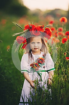 Beautiful little girl with a wreath of poppies in a white dress and collects a bouquet of wildflowers. cute child in poppy field