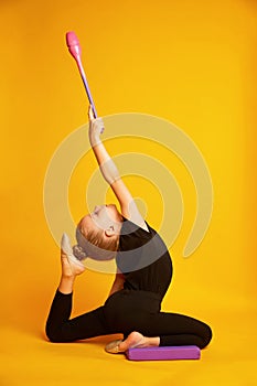 Beautiful little gymnast in black sportswear with gymnastic maces shows exercises on a yellow background.