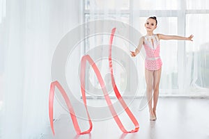 Beautiful little gymnast girl in pink sportswear dress, doing rhythmic gymnastics exercise Spirals with art ribbon