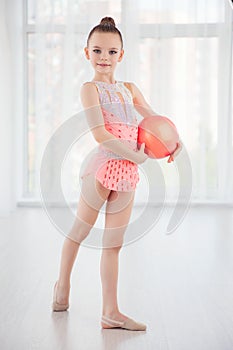Beautiful little gymnast girl in pink sportswear dress, performing art gymnastics element with ball in fitness class