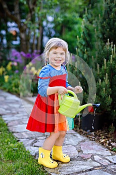 Beautiful little toddler girl in yellow rubber boots and colorful dress watering spring flowers with kids water can