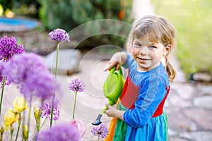 Beautiful little toddler girl in yellow rubber boots and colorful dress watering spring flowers with kids water can