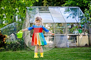 Beautiful little toddler girl in yellow rubber boots and colorful dress watering spring flowers with kids water can