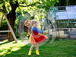 Beautiful little toddler girl in yellow rubber boots and colorful dress watering spring flowers with kids water can