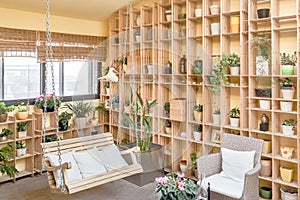 Beautiful living room interior with swing chair and cane sofa, stylish green plants and decorations on wooden shelves