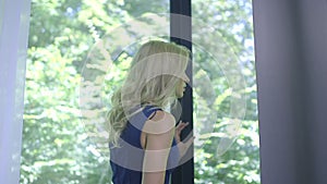 Beautiful lonely blonde woman standing at the window waiting for someone