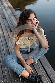 Beautiful lonely young curly girl in a yellow top with blue jeans sits on a wooden pier near the water