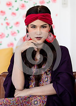 A beautiful long black hair lady with a red turban and purple blanket is sitting with elegant gesture on a big dark brown sofa in