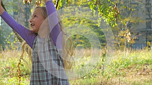 Beautiful long blonde hair girl holds yellow leaves on nature background. Smiling child plays in autumn park sunny day