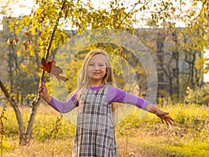 Beautiful long blonde hair girl holds yellow leaves on nature background. Smiling child plays in autumn park sunny day