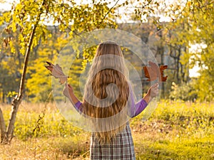 Beautiful long blonde hair girl holds yellow leaves on nature background. Smiling child plays in autumn park sunny day